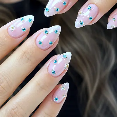 Close-up of a hand with almond nails in pale pink and white, set with blue gemstones and pearl flowers.