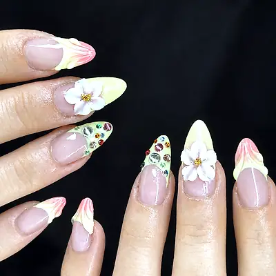 Two hands with pale yellow and pink gel nails decorated with 3D white flowers and coloured rhinestones.